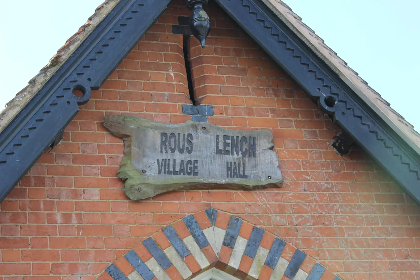 Wooden sign reading 'Rous Lench Village Hall' on a red brick gable with decorative ironwork